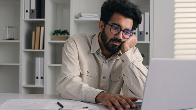 Arabian Indian ethnic male tired businessman office employee boring work with laptop dreary exhausted lazy bored business man low energy overworked fatigue depression apathy burnout typing on computer