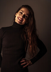 Enjoying toothy smiling makeup woman with brown long hair posing in black sweater on dark shadow background. Closeup