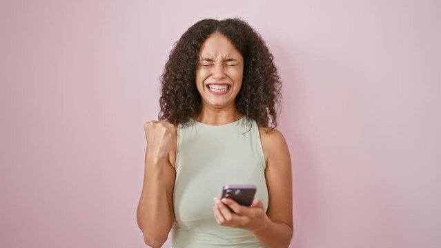 Cheerful young hispanic woman with curly hair joyfully celebrating messaging win over pink isolated background, happily using smartphone displaying winner gesture