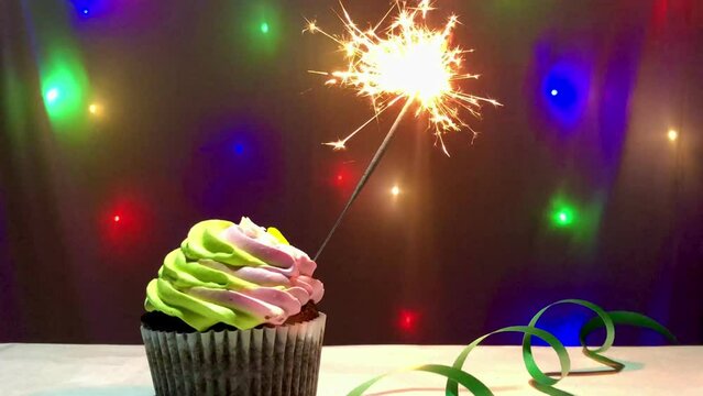 Birthday cupcake with sparkler and multicolor lights on the background and green Serpentine on the table