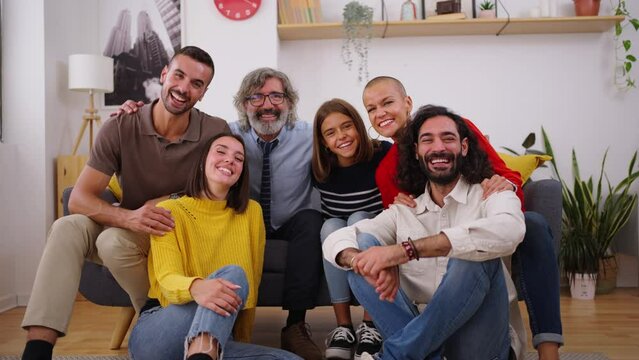 Portrait of happy Caucasian family posing smiling looking at camera hugging indoors. Multi generational joyful people gathered around the sofa of living room for celebrating together at home