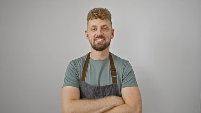 A young handsome caucasian man with blue eyes and a beard wearing a denim apron poses with crossed arms against an isolated white background.