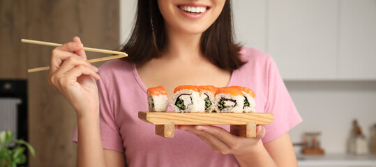 Young woman eating tasty sushi rolls in kitchen - obrazy, fototapety, plakaty