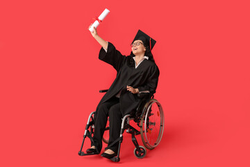 Beautiful female graduate student in wheelchair with graduation cap and diploma on red background