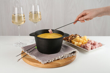 Dipping piece of ham into fondue pot with tasty melted cheese at white table, closeup