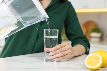 Woman pouring water from filter jug into glass in kitchen, closeup