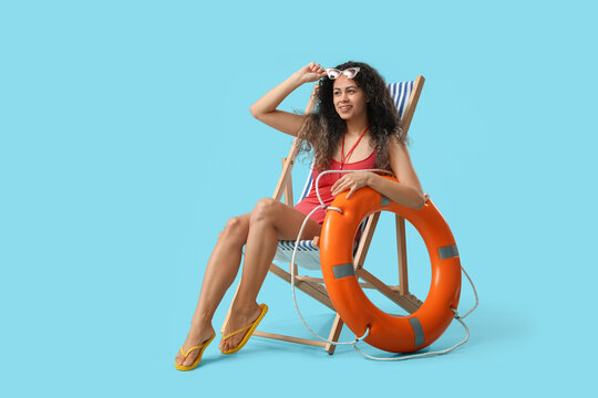 Beautiful young happy African-American female lifeguard with ring buoy sitting on deckchair against blue background