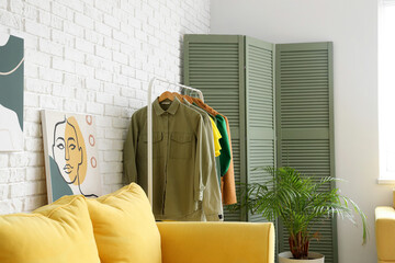 Rack with female clothes, houseplant and folding screen near light brick wall in room