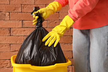 Woman taking full garbage bag from trash bin near brick wall