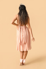 Beautiful young African-American woman in stylish prom dress and tiara on brown background, back view