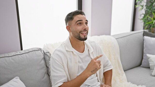 Young happy arab man with a smart face, sitting on his indoor sofa at home, smiles while cheerfully pointing his finger up, gesturing number one, with a successful idea and sheer excitement.