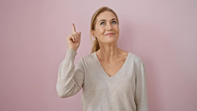 Cheerful middle age blonde woman has a light bulb moment! standing, pointing finger up signifying number one, she's brimming with success. isolated over pink wall, she's one happy thinker!