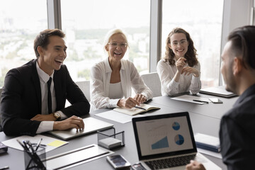 Happy professional team members negotiate in modern office, joking, laughing, take part in briefing event to share opinion and creative ideas, sit at desk enjoy friendly talk and successful teamwork