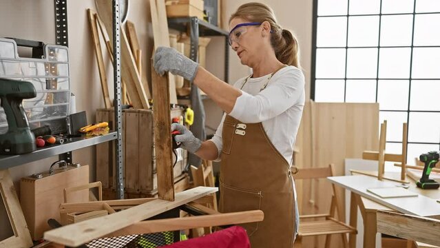 Mature woman wearing safety glasses and apron planes wood in a well-lit carpentry workshop