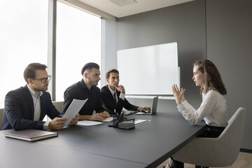 Young attractive woman vacancy candidature passing job interview in company office boardroom, three male HR managers asking questions, evaluating female applicant sit at table in meeting room. Hiring