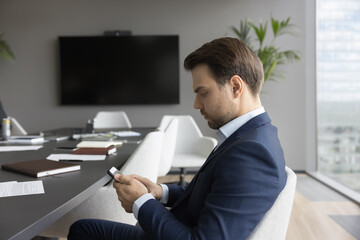 Serious young businessman sit in boardroom, type formal message use smartphone, download new mobile...