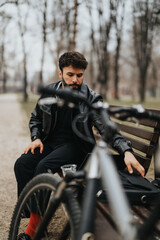 Focused businessman in stylish leather jacket uses laptop while sitting next to his bicycle in an urban park setting.