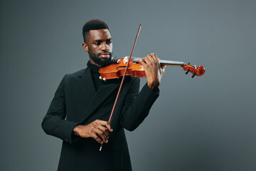 Elegant African American male musician in black suit playing violin against gray background,...