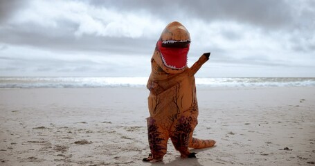 Mascot, funny and dinosaur costume on beach dancing with energy for comedy joke on vacation travel. Goofy, silly and inflatable animal outfit moving, jumping and having fun by ocean on holiday.