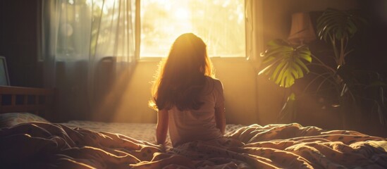A woman is quietly seated on a bed, looking out towards a bright window in a serene room - obrazy, fototapety, plakaty