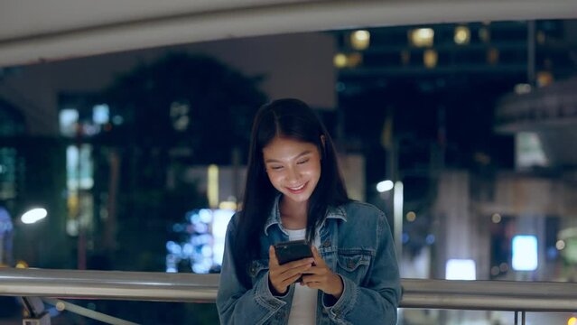Beautiful young Asian woman standing in city street at night using mobile smartphone. Happy woman standing and typing messages on her mobile phone