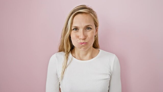Cheeky blonde gal pulls a hilarious face, puffs cheeks full of air over pink isolated background