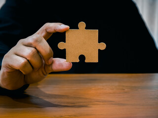 A jigsaw puzzle in businessman hand. Man in black shirt showing empty piece of important jigsaw puzzle on wood desk. Business solution and idea, partnership, elements of career success part concept.