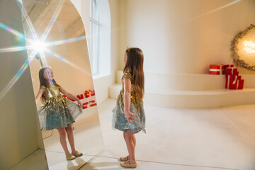 Beautiful little girl in a shiny dress looking in the mirror at her reflection. 
