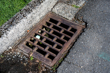 Closeup of a rusty iron storm drain grate in a street with a pvc pipe feeding into it
