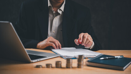 Man in a suit stamp approves documents at a wooden desk with a laptop and coins. business finance...