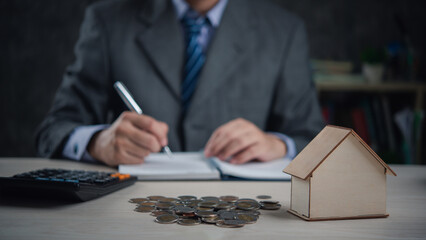 Agent in suit takes notes by house model and coin stacks, suggesting real estate investment or writing contract and insurance buy and sell home. business finance concept - 783506859
