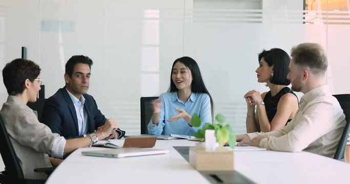 Positive beautiful young Asian team leader woman talking to multiethnic colleagues, explaining creative idea to diverse team, speaking at large meeting table, enjoying teamwork communication