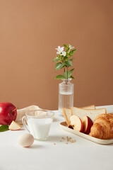 Layout of the meal for breakfast or brunch with a cup of milk, some apple pieces, a yummy croissant and sandwiches, a transparent vase of flower on basic brown background. 