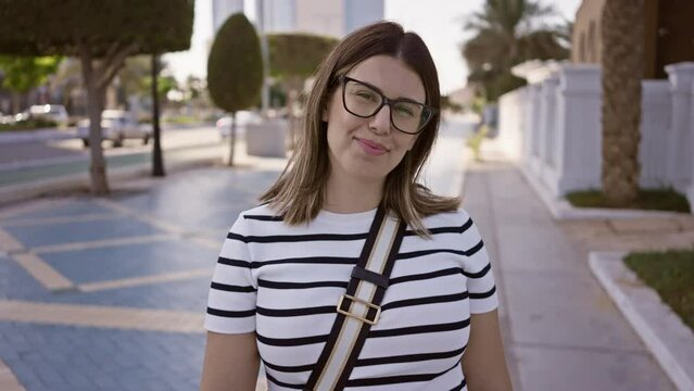 A smiling woman wearing glasses stands on an urban street in abu dhabi, with modern cityscape in the background.