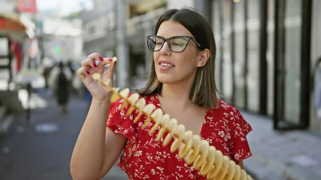 Beautiful hispanic woman joyfully eating crunchy, delicious chips on a stick at takeshita street, tokyo - traveling junk food lover, sporting glasses and a cheerful smile in japan's urban city