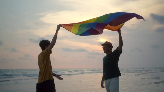 Romantic moment of bisexual gay male couple walking on the beach with their arms crossed with rainbow flag. LGBTQ man couple have a romantic moment on the beach. LGBT and homosexual concept.