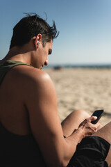 Young man using wireless earbuds and smartphone on beach in sunlight