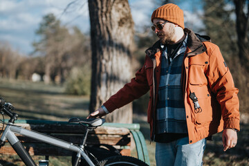 Fototapeta premium Outdoor portrait of a fashionable man wearing a warm autumn jacket and beanie standing next to a bicycle in a park.