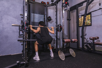 Fototapeta na wymiar Dedicated and focused novice male doing back squats facing the wall on a smith machine in a well-equipped gym. A beginner starting to workout and train.