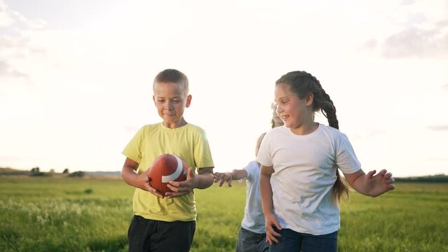 Group of children with rugby ball play American football. In summer, team of kid in nature plays with an american football rugby ball. Kid run in park at sunset. Team of children together in nature