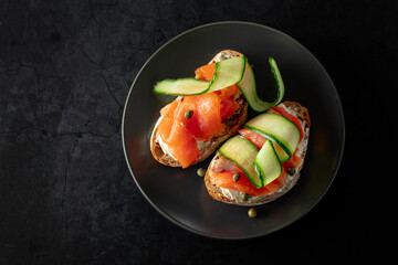 Sandwiches with smoked trout, cream cheese, fresh cucumber, and capers on a black plate. - 783597031