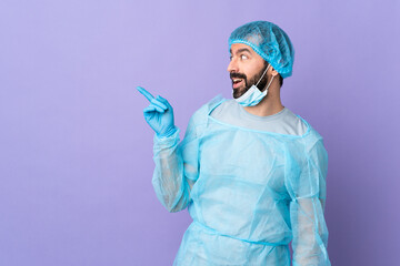 Surgeon man with beard with blue uniform over isolated purple background intending to realizes the solution while lifting a finger up - obrazy, fototapety, plakaty
