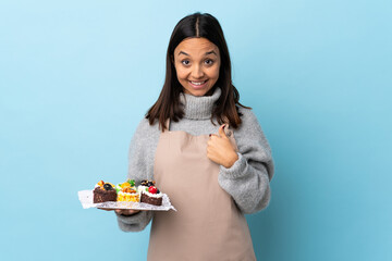 Pastry chef holding a big cake over isolated blue background with surprise facial expression.