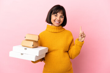 Pregnant woman holding pizzas and burgers isolated on pink background pointing up a great idea