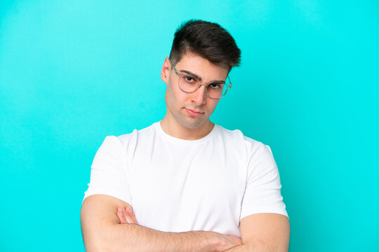 Young caucasian man isolated on blue background With glasses and arms crossed