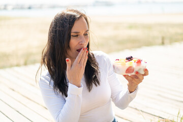 Middle aged woman holding a bowl of fruit at outdoors with surprise and shocked facial expression