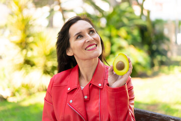 Middle aged woman holding an avocado at outdoors looking up while smiling