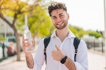Young handsome man with a bottle of water at outdoors with thumbs up because something good has happened