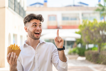 Young handsome man holding fried chips at outdoors intending to realizes the solution while lifting a finger up - obrazy, fototapety, plakaty