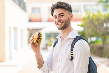 Young handsome man holding a burger at outdoors smiling a lot